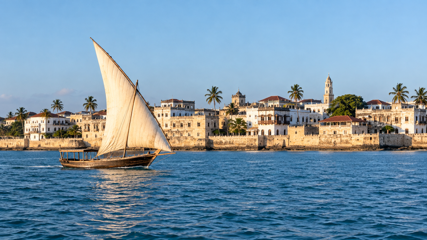 Un dhow de vela clara cruza el agua tranquila frente al paseo marítimo de Stone Town.